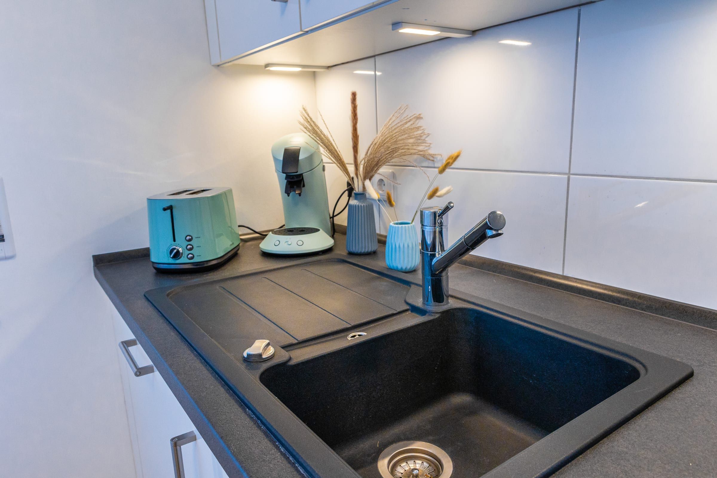Spüle aus dunklem Granit, dahinter Toaster und Kaffeemaschine unter der Arbeitsplattenbeleuchtung