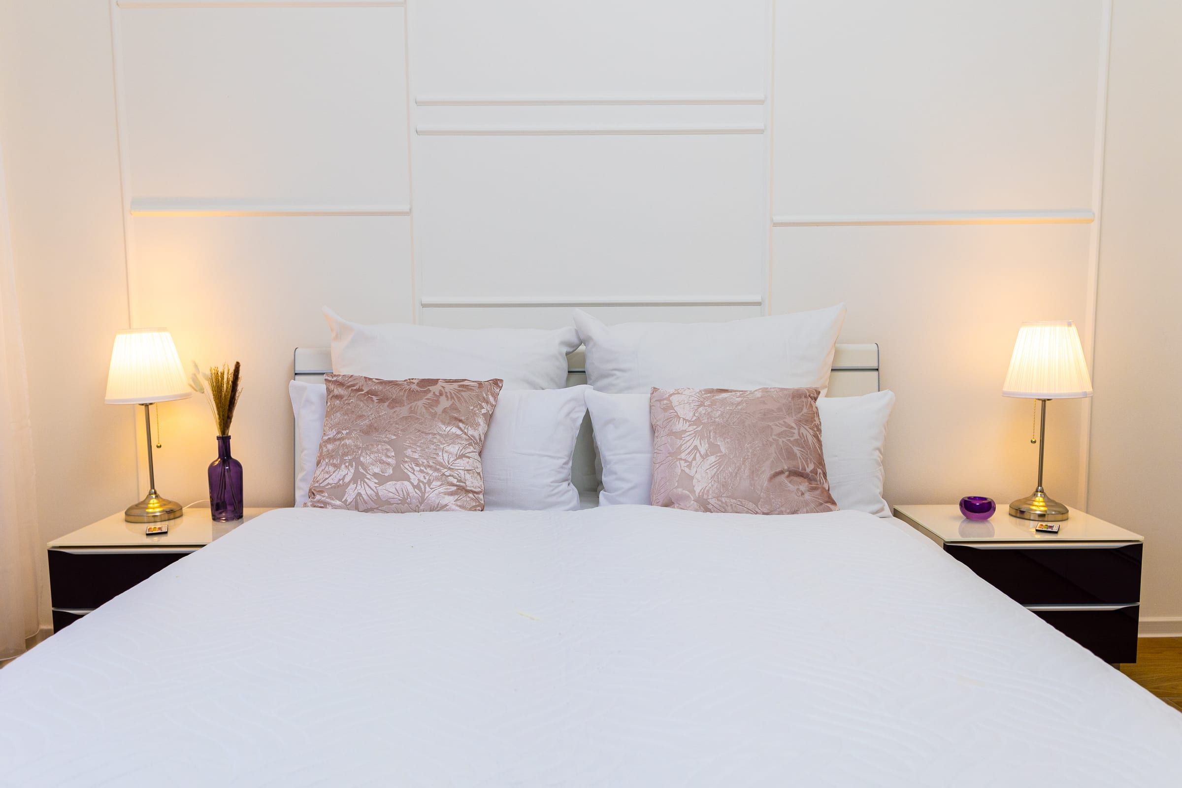 Doppelbett mit weißer Tagesdecke, rosa Kissen und zwei Nachttischlampen