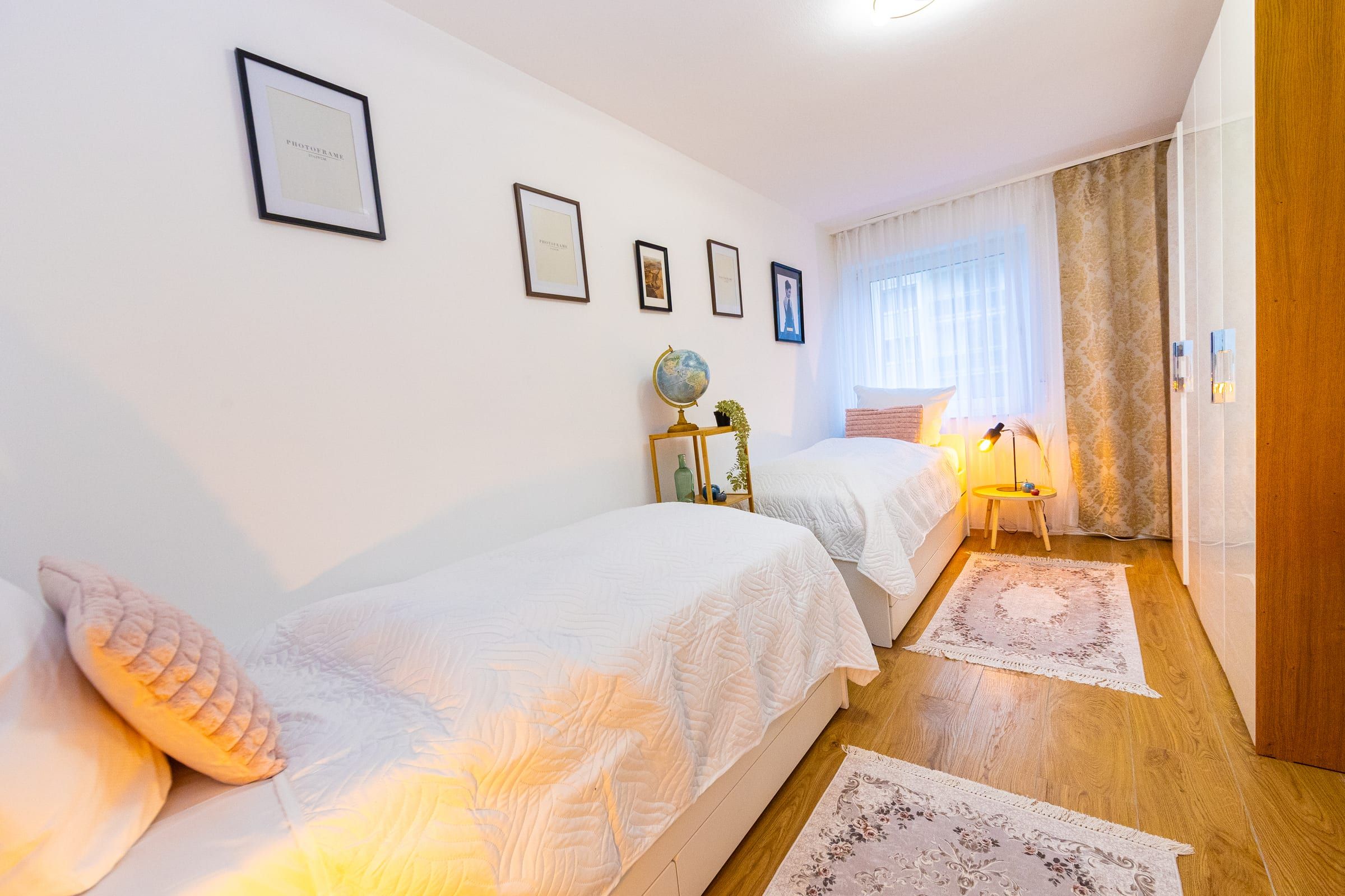 Einzelbett mit weißer Tagesdecke und rosa Kissen, im Hintergrund das Fenster mit Vorhang