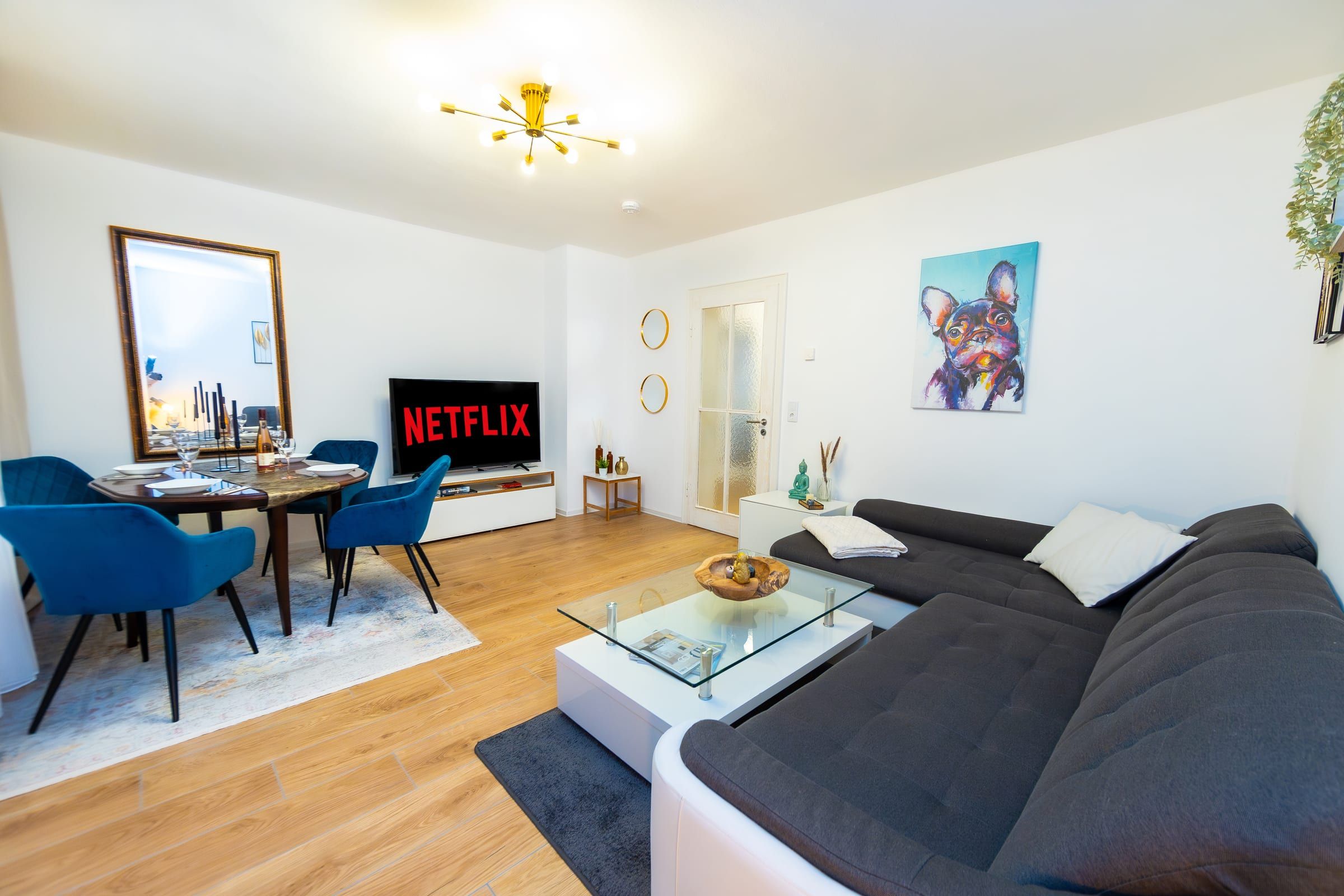 Helles Wohnzimmer mit dunkelgrauem Ecksofa, Couchtisch aus Glas und Esstisch mit blauen Samtstühlen