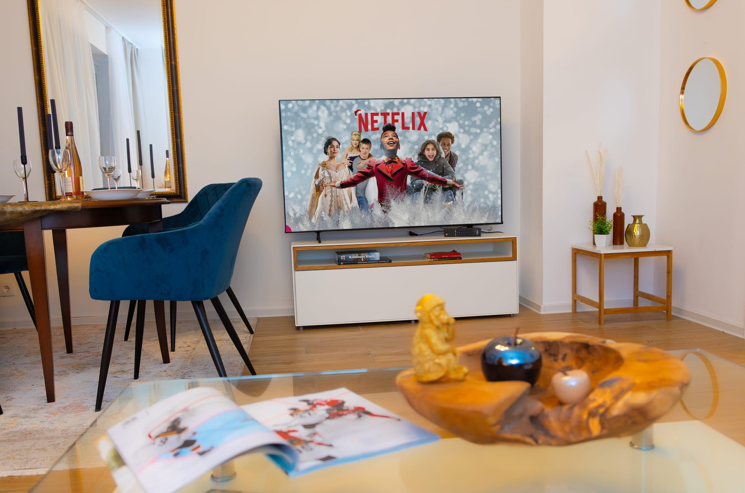 Fernseher auf weißem Lowboard, links der Esstisch mit blauen Stühlen, vorne ein Glastisch mit Holzschale