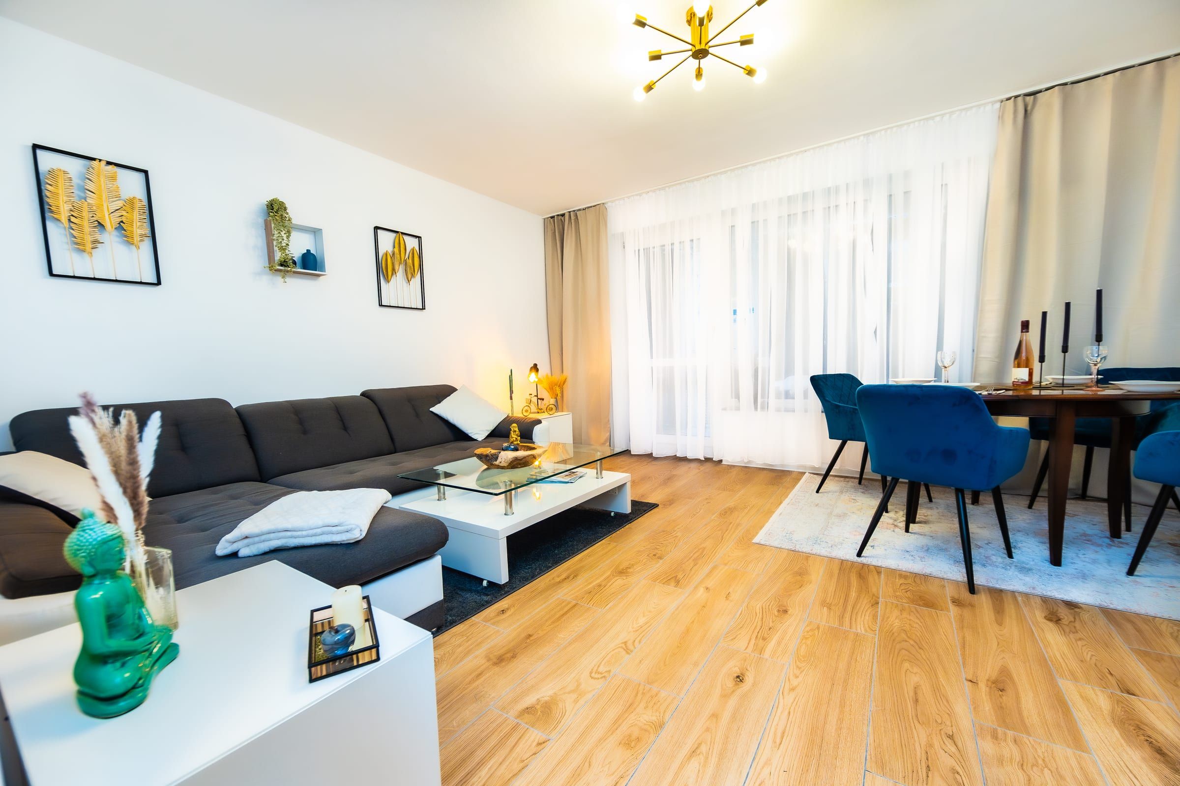 Wohnzimmer mit dunkelgrauem Ecksofa und weißem Couchtisch, dahinter Esstisch und große Fensterfront mit Vorhängen